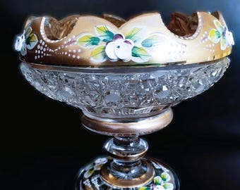 May include: A clear, cut-glass pedestal bowl with a gold-painted rim and base. The rim features a floral design with white flowers, green leaves, and gold accents. The bowl is likely used for decorative purposes.