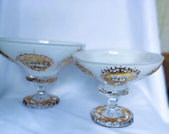 May include: Two elegant, white glass bowls with gold and floral accents. The bowls have a wide, shallow design and sit on ornate, clear glass stems with gold detailing. A decorative piece for serving.