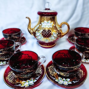 Op de afbeelding: Een robijnrode theeset met gouden accenten, bestaande uit een theepot en zes kopjes met schotels. De theepot is goudkleurig met bloemen- en hartmotieven. De kopjes en schotels hebben gouden randen en decoratieve patronen.