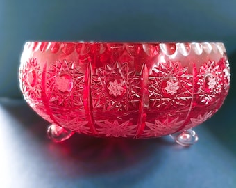 May include: A ruby red, cut glass bowl with a scalloped rim and starburst and geometric patterns. The bowl rests on three small, clear feet. The bowl is likely used for decorative purposes or serving.