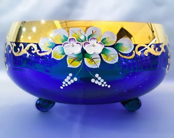 May include: A cobalt blue glass bowl with gold trim and hand-painted floral designs. The bowl has three feet and is perfect for serving fruit or snacks.