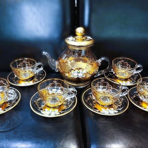 Puede incluir: Juego de té de cristal transparente con detalles dorados. El juego incluye una tetera con tapa dorada y seis tazas con platillos a juego. Los platillos y la tetera presentan diseños florales dorados.
