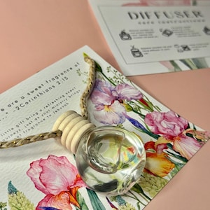 May include: A clear glass diffuser with a wooden top and a braided cord, resting on a floral card with pink and purple irises. The card includes text from 2 Corinthians 2:15. A second card with instructions is in the background.