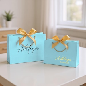 Pode incluir: Duas sacolas de presente azul claro com laços dourados e alças de corda. A sacola maior tem o nome "Ashleya" em escrita preta. A sacola menor tem "Ashleya" em escrita dourada com "Bridesmaid" abaixo.