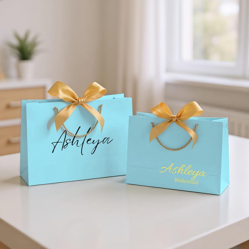 May include: Two light blue gift bags with gold bows and rope handles. The larger bag has the name "Ashleya" in black script. The smaller bag has "Ashleya" in gold script with "Bridesmaid" below.