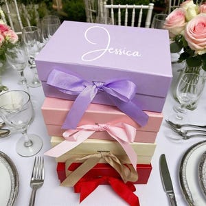 Puede incluir: Una pila de cajas de regalo en varios colores: lavanda, rosa, crema y rojo. Cada caja está atada con una cinta a juego. La caja superior es de color lavanda y tiene el nombre "Jessica" en escritura blanca.