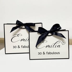 May include: Two white gift bags with black trim and black satin bows. The bags feature the name "Emilia" in cursive script, and the phrase "30 & fabulous" printed in a simple font.