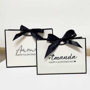 May include: Two white gift bags with black trim and black satin bows. The bags have the name "Amanda" printed in black script, along with "HAPPY VALENTINES" or "HAPPY VALENTINES DAY" in black text. One bag has a small black heart.