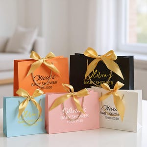 Peut inclure: Plusieurs sacs cadeaux color&eacute;s, orn&eacute;s de n&oelig;uds en satin dor&eacute;. Les sacs sont orange, noir, bleu clair, rose et blanc. Chaque sac porte une anse dor&eacute;e et l'inscription "Olivia's BABY SHOWER 10.08.2030".