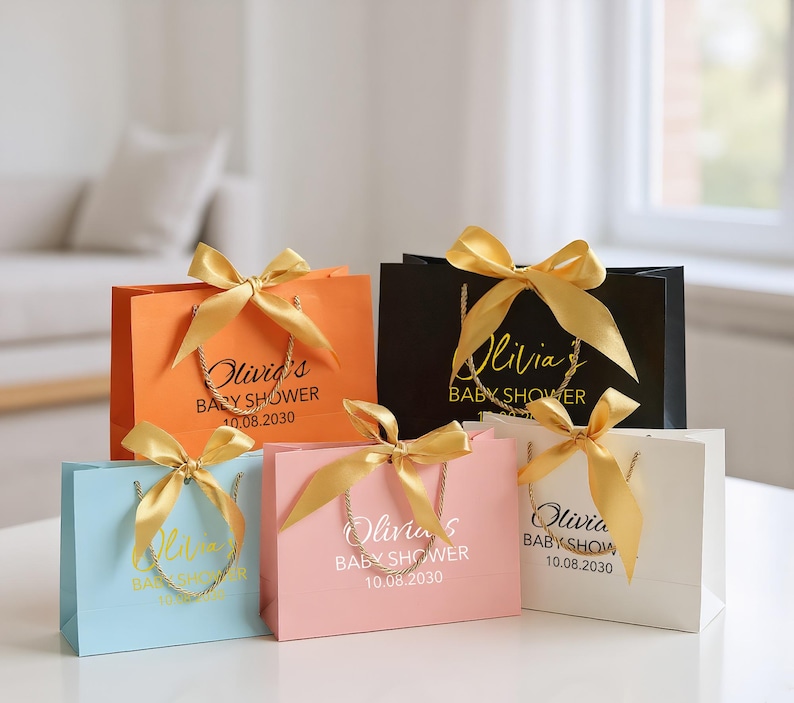 Peut inclure: Plusieurs sacs cadeaux color&eacute;s, orn&eacute;s de n&oelig;uds en satin dor&eacute;. Les sacs sont orange, noir, bleu clair, rose et blanc. Chaque sac porte une anse dor&eacute;e et l'inscription "Olivia's BABY SHOWER 10.08.2030".