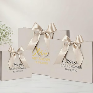 Peut inclure: Trois sacs cadeaux gris clair de diff&eacute;rentes tailles, chacun orn&eacute; d'un n&oelig;ud en ruban de satin. Les sacs ont des poign&eacute;es en corde et pr&eacute;sentent le texte "Olivia BABY SHOWER 10.08.2030" en or et noir. Un vase de fleurs blanches est en arri&egrave;re-plan.