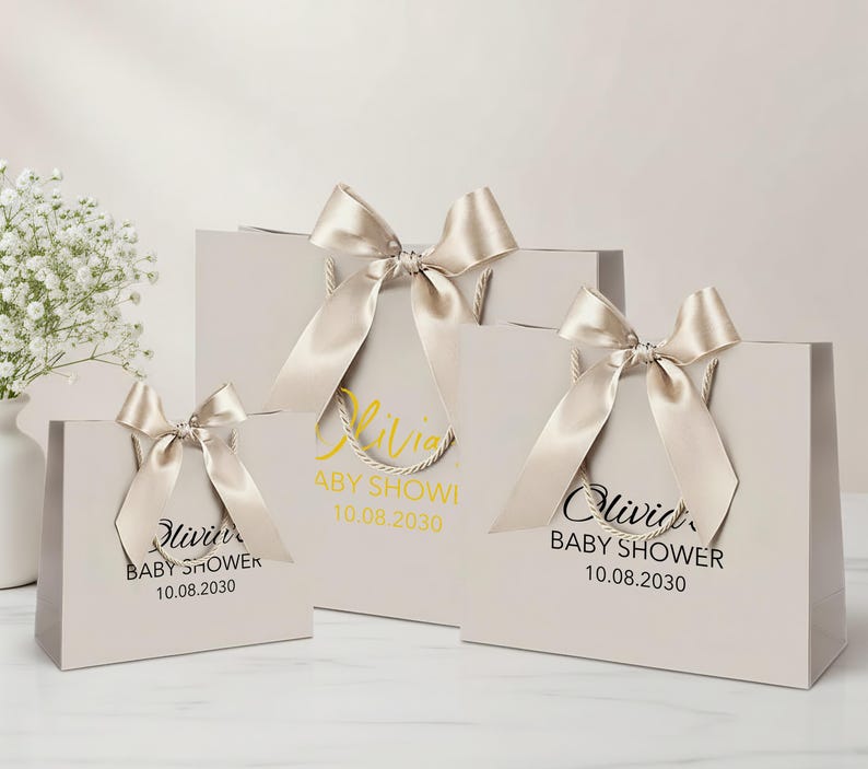 Peut inclure: Trois sacs cadeaux gris clair de diff&eacute;rentes tailles, chacun orn&eacute; d'un n&oelig;ud en ruban de satin. Les sacs ont des poign&eacute;es en corde et pr&eacute;sentent le texte "Olivia BABY SHOWER 10.08.2030" en or et noir. Un vase de fleurs blanches est en arri&egrave;re-plan.