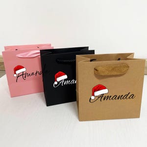 May include: Three gift bags in pink, black, and brown. Each bag features a Santa hat graphic and the name "Amanda" in script. The pink bag has a pink ribbon handle, the black bag has a black cord handle, and the brown bag has a woven handle.