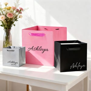 May include: Three square gift bags in pink, black, and silver. The large pink bag features the name Ashleya in black script. The black bag features the name Ashleya in white script. The silver bag features the name Ashleya and the word Bridesmaid.