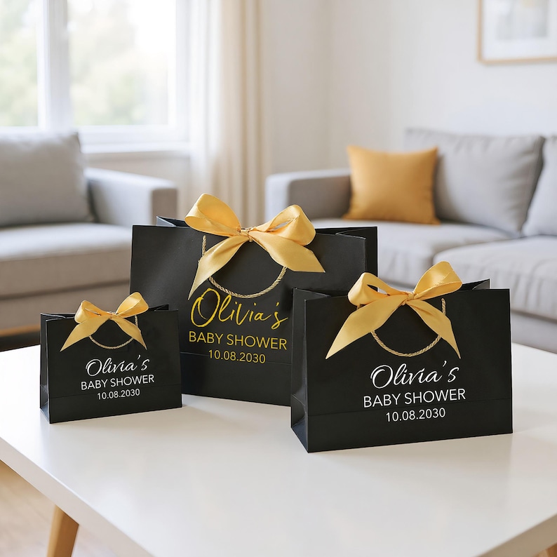 Peut inclure: Trois sacs cadeaux noirs de diff&eacute;rentes tailles, orn&eacute;s de n&oelig;uds dor&eacute;s. Les sacs portent l'inscription "Olivia's BABY SHOWER 10.08.2030" en or et blanc. Ils sont pos&eacute;s sur une table blanche.