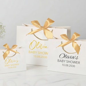 Peut inclure: Trois sacs cadeaux blancs de diff&eacute;rentes tailles, chacun orn&eacute; d'un n&oelig;ud en satin dor&eacute;. Les sacs pr&eacute;sentent le texte "Olivia's BABY SHOWER 10.08.2030" en lettres dor&eacute;es et noires, avec des poign&eacute;es en corde dor&eacute;e.