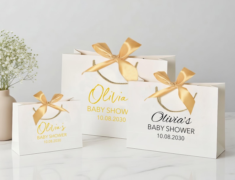 Peut inclure: Trois sacs cadeaux blancs de diff&eacute;rentes tailles, chacun orn&eacute; d'un n&oelig;ud en satin dor&eacute;. Les sacs pr&eacute;sentent le texte "Olivia's BABY SHOWER 10.08.2030" en lettres dor&eacute;es et noires, avec des poign&eacute;es en corde dor&eacute;e.