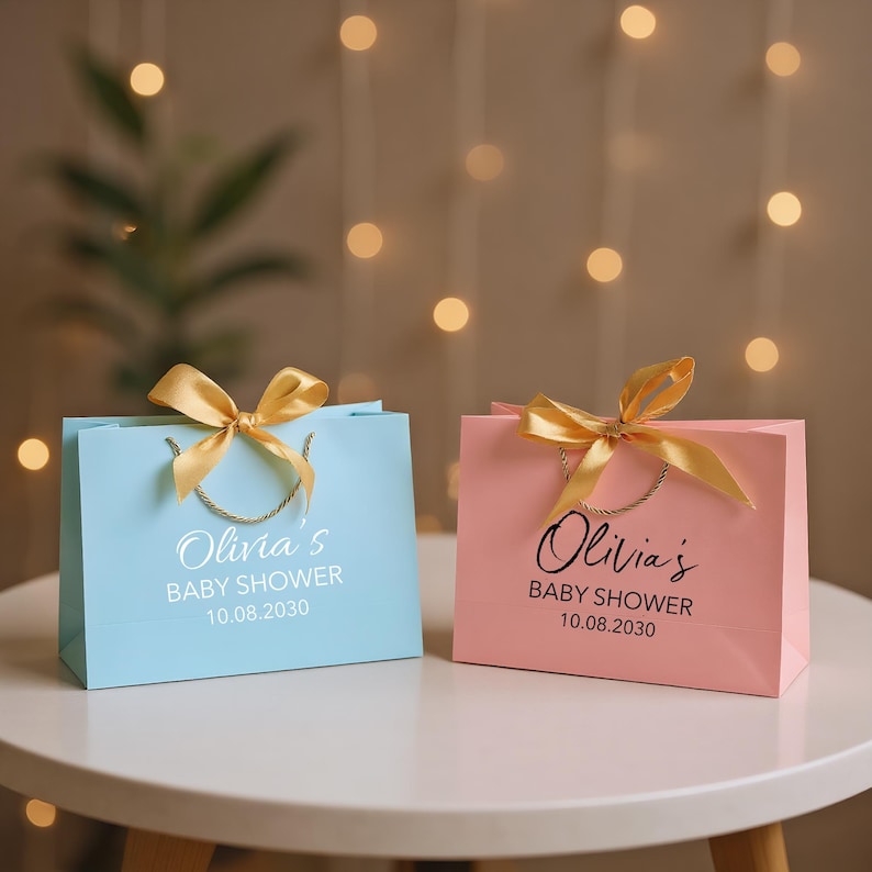 Peut inclure: Deux sacs cadeaux, un bleu clair et un rose, pos&eacute;s sur une table blanche. Chaque sac a un n&oelig;ud en ruban dor&eacute; et des poign&eacute;es en corde dor&eacute;e. Les sacs portent l'inscription "Olivia's BABY SHOWER 10.08.2030" en &eacute;criture blanche et noire.