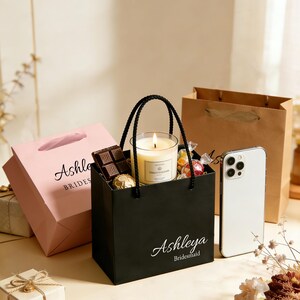 May include: A black gift bag with the text "Ashleya Bridesmaid" contains a lit candle, chocolates, and wrapped candies. A pink gift bag with "Ashley Brides" is to the left. A white smartphone and a brown paper bag are also present.