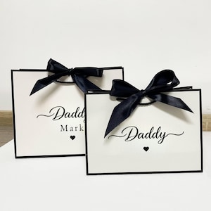 Puede incluir: Dos bolsas de regalo blancas con ribete negro y lazos de satén negro. Las bolsas tienen la palabra "Daddy" impresa en una fuente cursiva, con un símbolo de corazón. Una bolsa también tiene el nombre "Mark" impreso debajo de "Daddy".
