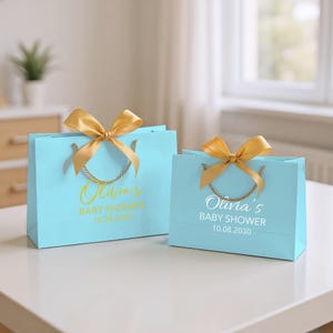 Peut inclure: Deux sacs cadeaux bleu clair avec des n&oelig;uds et des poign&eacute;es dor&eacute;s. Les sacs portent l'inscription "Olivia's BABY SHOWER 10.08.2030" en lettres dor&eacute;es et blanches. Les sacs sont sur une surface blanche.