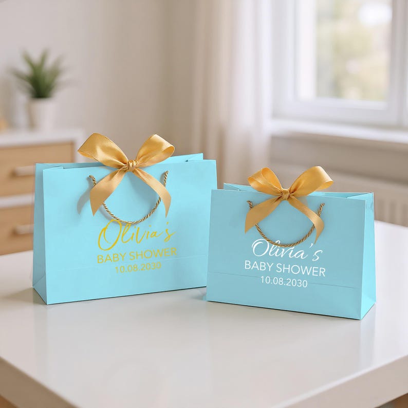 Peut inclure: Deux sacs cadeaux bleu clair avec des n&oelig;uds et des poign&eacute;es dor&eacute;s. Les sacs portent l'inscription "Olivia's BABY SHOWER 10.08.2030" en lettres dor&eacute;es et blanches. Les sacs sont sur une surface blanche.