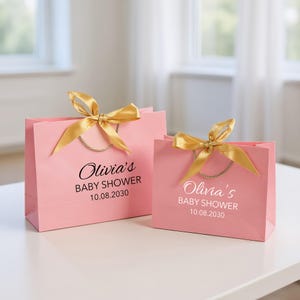 Peut inclure: Deux sacs cadeaux roses avec des n&oelig;uds en ruban dor&eacute; et des poign&eacute;es en corde. Les sacs portent l'inscription "Olivia's BABY SHOWER 10.08.2030" en noir. Les sacs sont sur une surface blanche.