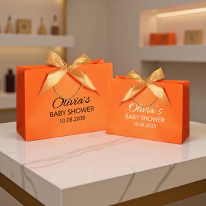 Peut inclure: Deux sacs-cadeaux orange avec des n&oelig;uds et des poign&eacute;es en ruban dor&eacute;. Les sacs portent l'inscription "Olivia's BABY SHOWER 10.08.2030" en blanc. Les sacs sont pos&eacute;s sur une surface en marbre blanc.