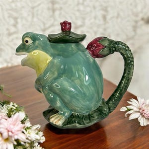 Puede incluir: Una tetera de cerámica caprichosa con forma de rana, en tonos verdes y amarillos. La tetera presenta un asa de capullo de flor roja y una tapa a juego. La rana está sentada sobre una base de hoja de nenúfar, lo que añade al diseño decorativo.