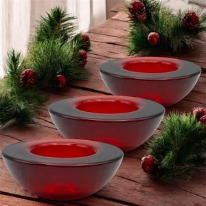 Op de afbeelding: Drie rode glazen kandelaars met een donkere rand. De kandelaars zijn rond met een ondiepe komvorm. De achtergrond bevat dennentakken en dennenappels, wat wijst op een vakantiethema.