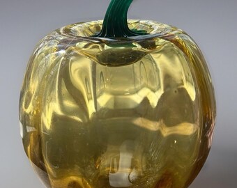 May include: A glass pumpkin with a green stem. The pumpkin is yellow and has a textured surface.