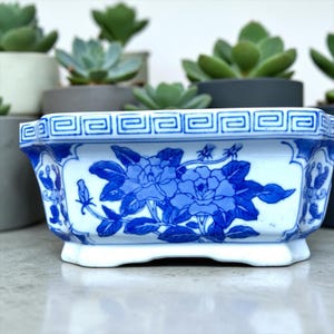 May include: A rectangular ceramic planter featuring a white base with blue floral patterns and a geometric Greek key border along the top rim. The planter has a flared base and is decorated with detailed blue blossoms and leaves on its side.