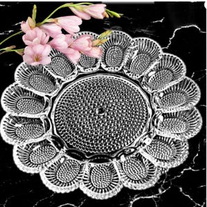 Puede incluir: Una fuente de vidrio transparente con un diseño floral, con un área circular central y compartimentos en forma de pétalo alrededor del borde. Flores rosas se colocan en la parte superior izquierda. La fuente está sobre una superficie negra.