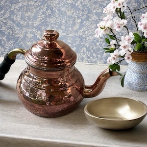 Könnte beinhalten: Eine Kupferteekanne mit gehämmerten Texturen, Messinggriff und schwarzem Griff steht neben einer kleinen, goldfarbenen Schale. Die Teekanne hat einen gebogenen Ausguss und einen gewölbten Deckel. Im Hintergrund steht eine Vase mit weißen Blumen.