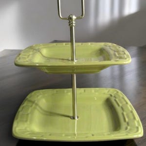 Puede incluir: Bandeja de servir de dos niveles en verde claro. Los niveles cuadrados tienen un borde decorativo y están conectados por un asa de metal dorado. Ideal para presentar pasteles o aperitivos.