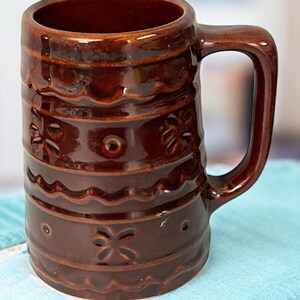 Könnte beinhalten: Ein brauner Keramikbecher mit Henkel. Der Becher hat eine glänzende Oberfläche und ist mit horizontalen Bändern und floralen Mustern verziert. Der Becher steht auf einem blauen Tuch.