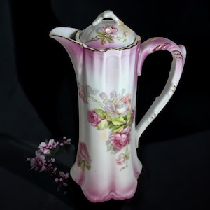 May include: A tall porcelain coffee pot featuring a white body with vertical fluted panels and a gradient pink rim. The surface is decorated with painted pink and white rose clusters, accented by gold trim on the handle, spout, and lid.