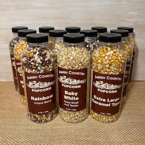 May include: A collection of clear plastic bottles filled with various types of popcorn kernels. The labels read "Amish Country Popcorn" with varieties like "Rainbow," "Baby White," and "Extra Large Caramel Type." Each bottle contains 14 oz of popcorn.