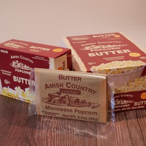 May include: Three boxes of Amish Country Butter Microwave Popcorn, with a single packet in front. The boxes are red and white, with the words "Butter" and "Popcorn" prominently displayed. The packet is brown with the brand name and product details.