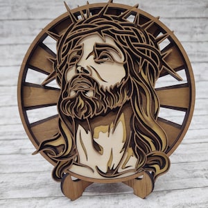 Op de afbeelding: Een gelaagd houten kunstwerk dat Jezus met een doornenkroon afbeeldt. De kleuren zijn bruin en beige, gemonteerd op een houten standaard. De achtergrond heeft een zonnestraalontwerp.