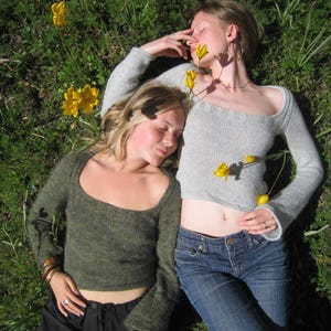 Könnte beinhalten: Zwei Personen in Pullovern und Jeans liegen in einem grasbewachsenen Feld mit gelben Blumen. Eine Person trägt einen grauen Pullover und blaue Jeans, die andere einen grünen Pullover und eine schwarze Hose. Die Pullover haben einen U-Boot-Ausschnitt.