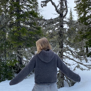 May include: A person wearing a gray hooded sweater and blue jeans standing in a snowy forest. The person is facing away from the camera.