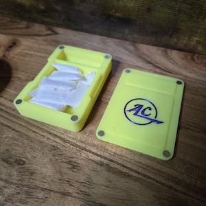 May include: A bright yellow rectangular box with a lid, open to reveal small white packets. The lid has a blue and silver logo that reads "AC". The box is on a wooden surface.