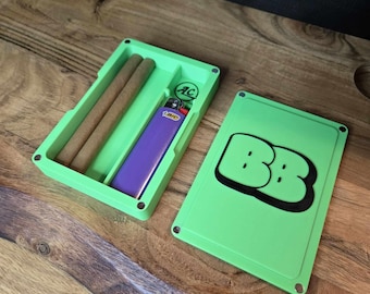 May include: A green, rectangular box with a lid. The box has a compartment for a lighter and two slots for rolling papers. The lid has a green and black logo that says "BB".