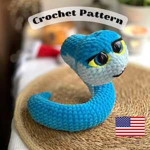 以下が含まれることがあります： 大きな黒い目と黄色のアクセントが付いた、手作りの青と白のかぎ針編みのヘビのおもちゃ。「Crochet Pattern」という文字がヘビの上にあります。おもちゃは織りマットの上にあります。アメリカの国旗が右下にあります。