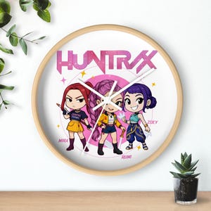May include: A round wall clock with a light wood frame. The clock face features a pink and white design with the word "HUNTRAX" and three cartoon characters. The characters are in the center, with the names "MIRA", "RUMI", and "ZOEY" below them.