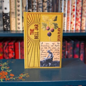 Op de afbeelding: Een hardcover boek genaamd "The Bell Jar" van Sylvia Plath. Het boek heeft een gele kaft met een vijgenillustratie en de titel in het rood. Een citaat en een zwart-witfoto staan op de kaft. Het boek ligt op een blauwe tafel.