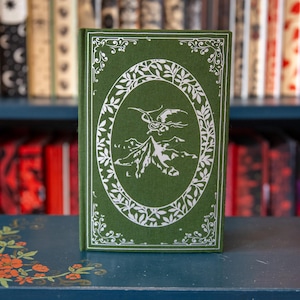 May include: A hardcover book with a green cover featuring a white oval design with a mountain and dragon illustration. The book is on a dark blue surface with floral accents, with a blurred background of other books on a shelf.