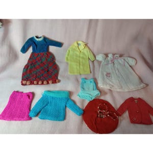 May include: A collection of doll clothing including a blue and plaid dress with pom-poms, a yellow coat, a white robe with a cat patch, a pink knit skirt, a blue knit sweater, a blue and white checkered romper, a red beret, and a red jacket.