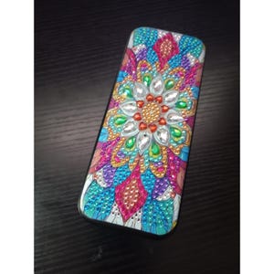 Puede incluir: Un estuche rectangular con un diseño de arte de diamantes de colores vibrantes. La superficie está cubierta con pequeños diamantes de imitación facetados en tonos rosa, azul, verde, morado y dorado, dispuestos en un patrón floral simétrico.